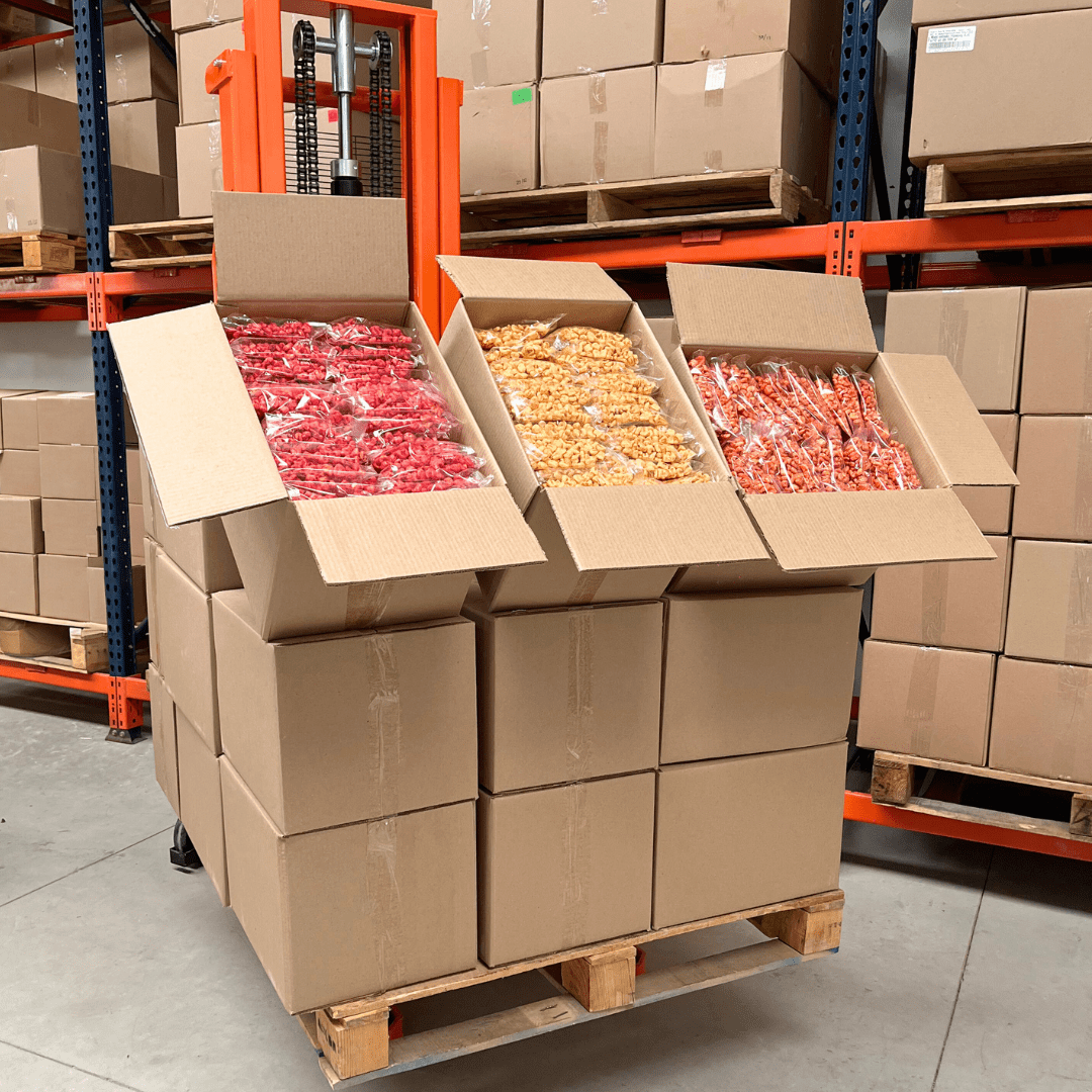 Palet con cajas de maicitos inflados al vapor en diferentes sabores listos para distribución al mayoreo. Fábrica y proveedor de botanas en México con inventario organizado para entrega a cafeterías escolares, gimnasios, universidades y tiendas naturistas. Venta al mayoreo de snacks como maicitos, obleas de amaranto, chips de vegetales y chicharrón de garbanzo, con opción de personalización de etiquetas para tu marca.