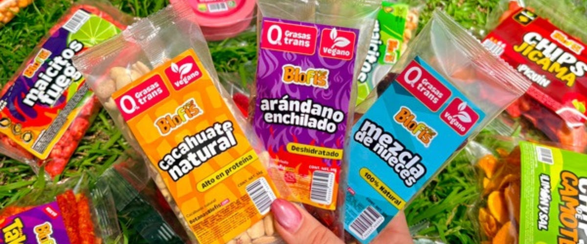 Mano con bolsas de botanas Blofis sobre el pasto: cacahuate natural, arándano enchilado y mezcla de nueces