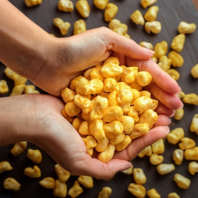 manos de mujer sosteniendo un puño de maicitos sabor jalapeño de fondo hay mas maicitos regados sobre la mesa de madera